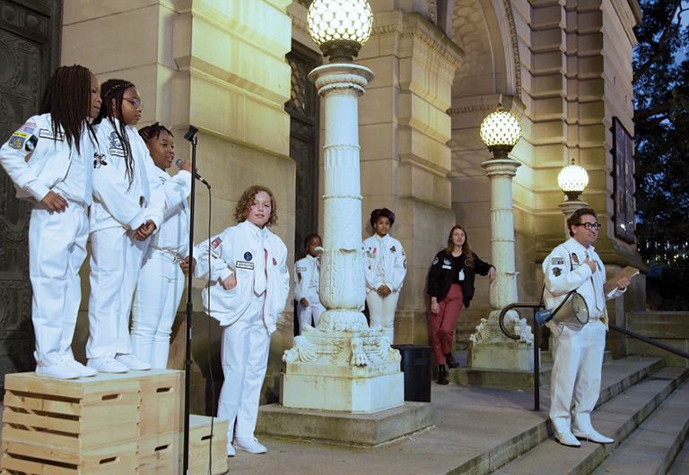 A group of children, dressed in white uniforms on the steps in front of the museum, with microphone stands in front of them.