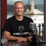 A portrait of Jason Brown sitting in a wet lab with a t-sheet with type saying science is greater than opinion.