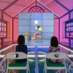Two young girls sitting in chairs inside a neon house watching a video.