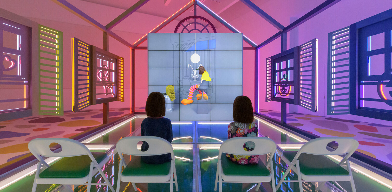 Two young girls sitting in chairs inside a neon house watching a video.