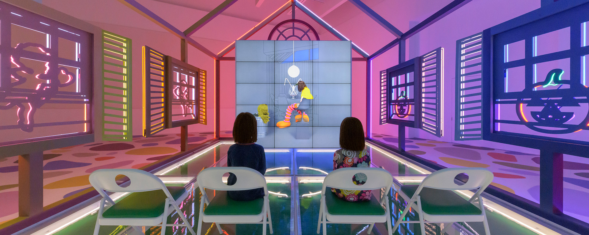 Two young girls sitting in chairs inside a neon house watching a video.