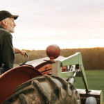 A farmer sittin gon a tractor looking over a field during sunset.