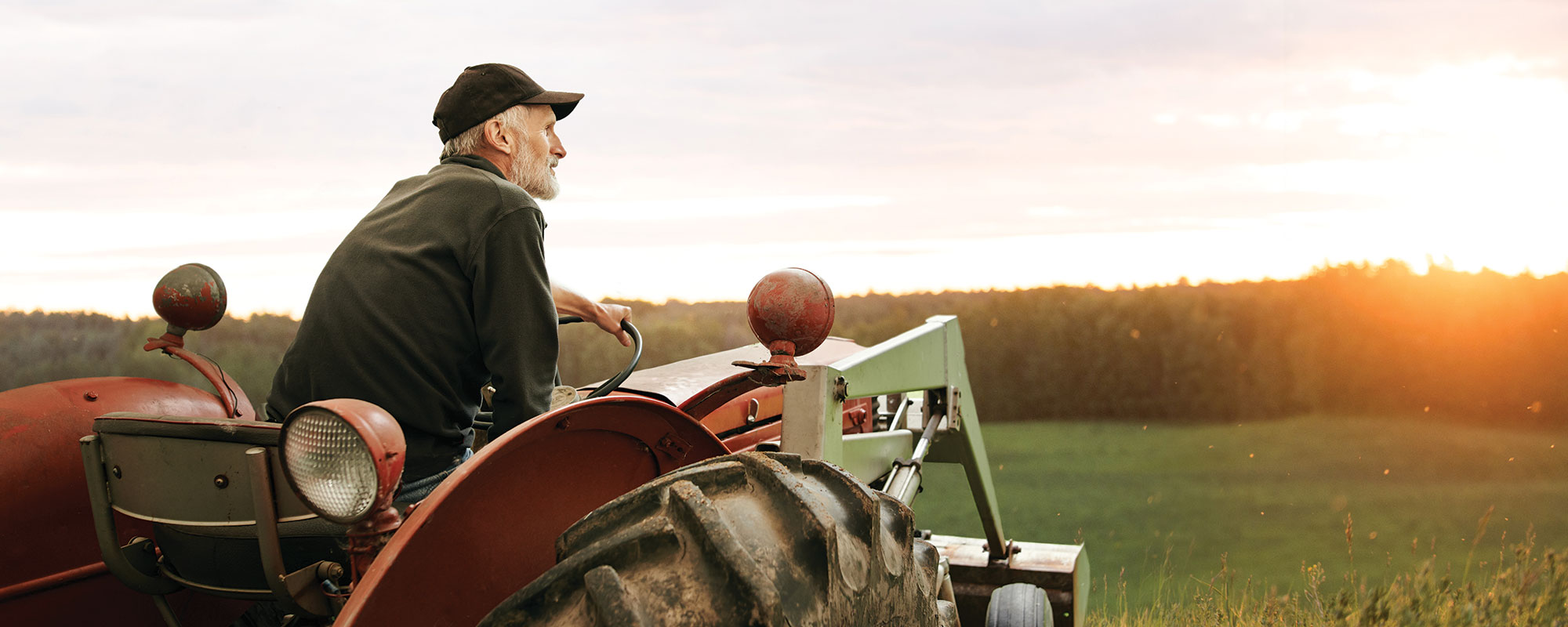 A farmer sittin gon a tractor looking over a field during sunset.