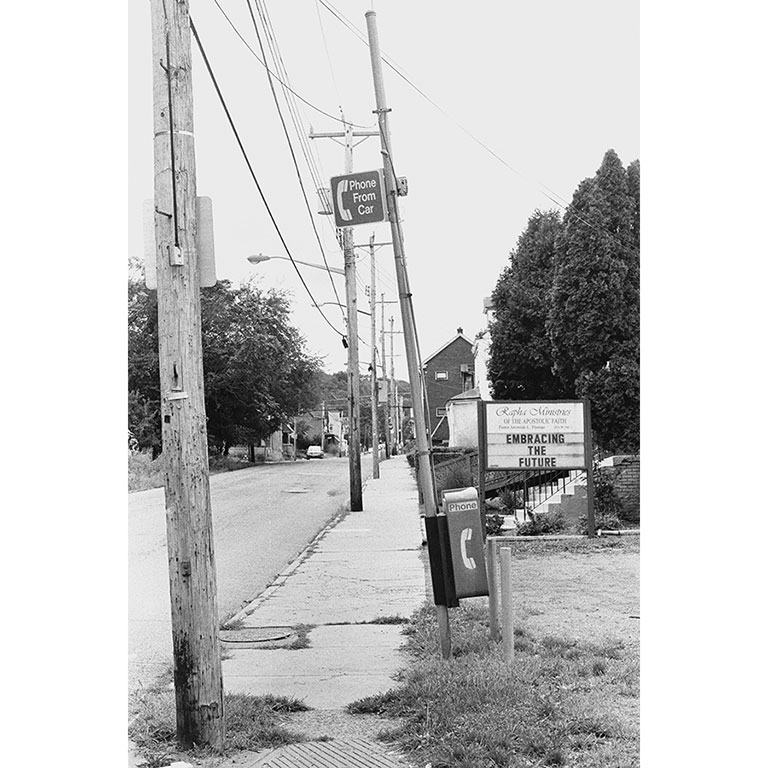 A black and white photo of a street, showing the sidewalk, a pay phone and a sign that says Embracing the Future.