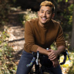 A photo of a young man in a brown shirt, sitting on a bike and smiling