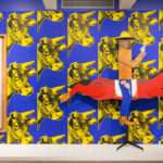 A boxy wooden sculture of teh actor, John Wayne holding a gun, on a horse in front of a wall of cow patterned wallpaper