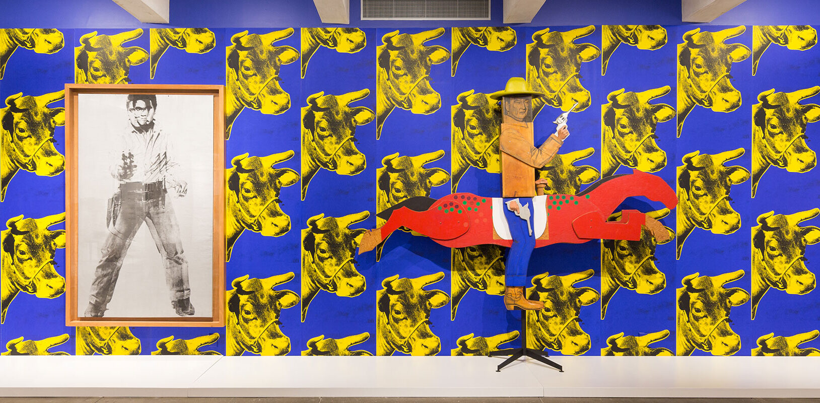 A boxy wooden sculture of teh actor, John Wayne holding a gun, on a horse in front of a wall of cow patterned wallpaper