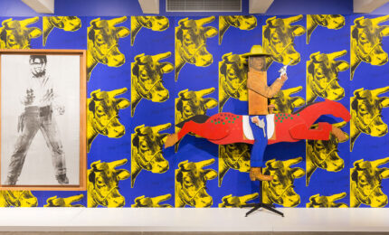A boxy wooden sculture of teh actor, John Wayne holding a gun, on a horse in front of a wall of cow patterned wallpaper