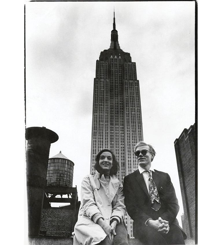A black and white photo of Andy Warhol and Marisol sitting with the empire state building behind them