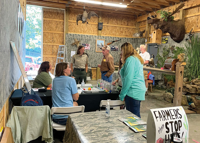 A group of people standing around a table talking to each other. A sign in the foreground reads, Farmers please stop here.