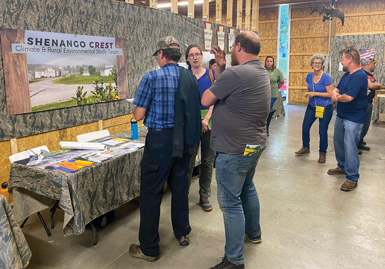 A group of people talking in front of a display at a fair. A sign reads Shenango Crest. Climate and Rural environmental study team.