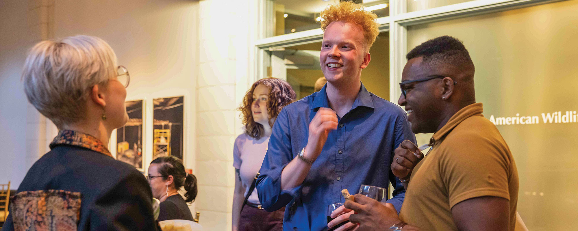 A group of three people engage in conversation at a social event, smiling and laughing, with others in the background.