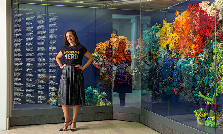 A woman in a "Museum Nerd" t-shirt and black skirt poses confidently in front of a vibrant, colorful art installation.