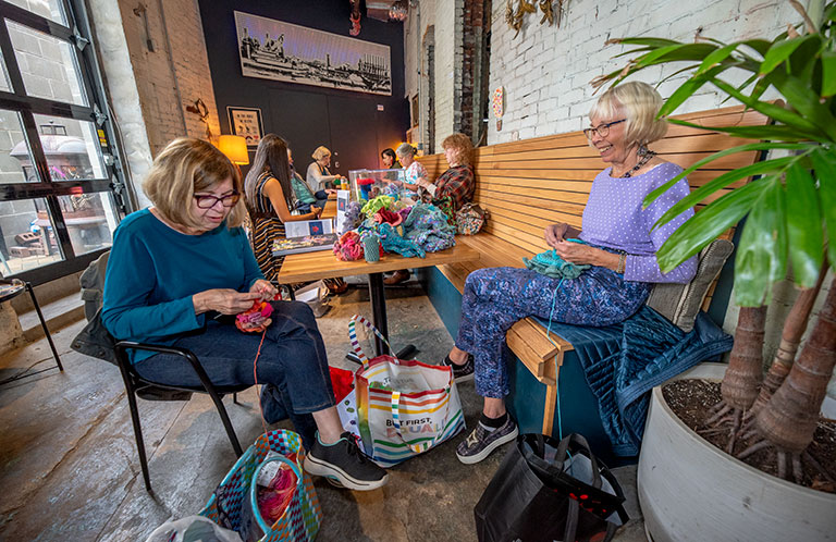 A cozy gathering of women knitting and chatting in a bright café with exposed brick walls and greenery around them.