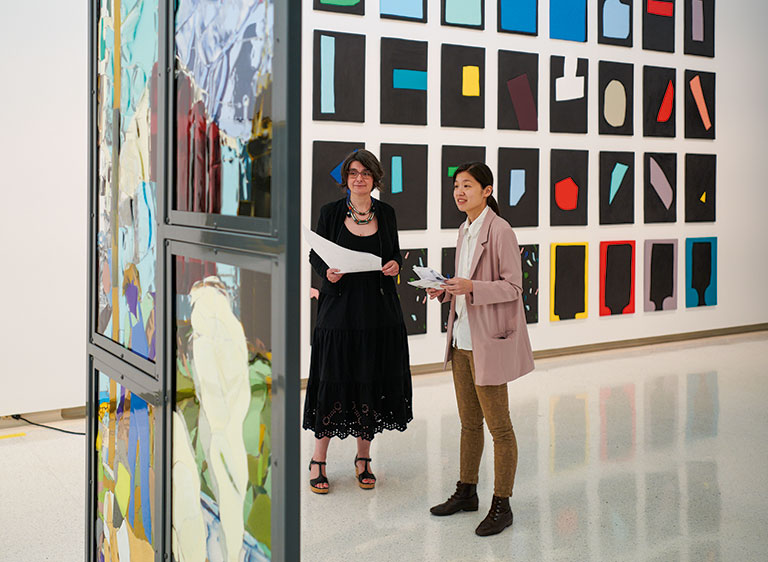 Two women converse in an art gallery, surrounded by colorful abstract artwork on the walls and reflective glass panels.