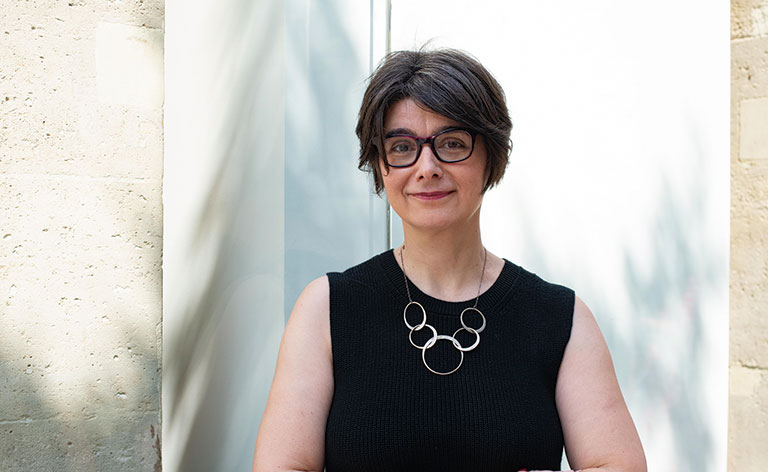 A woman with short gray hair and glasses stands confidently, wearing a black sleeveless top and circular necklace against a light background.