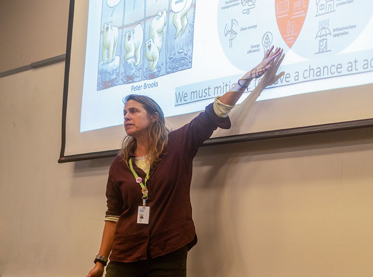 A woman gestures while presenting in front of a screen displaying cartoons and text about climate action strategies.