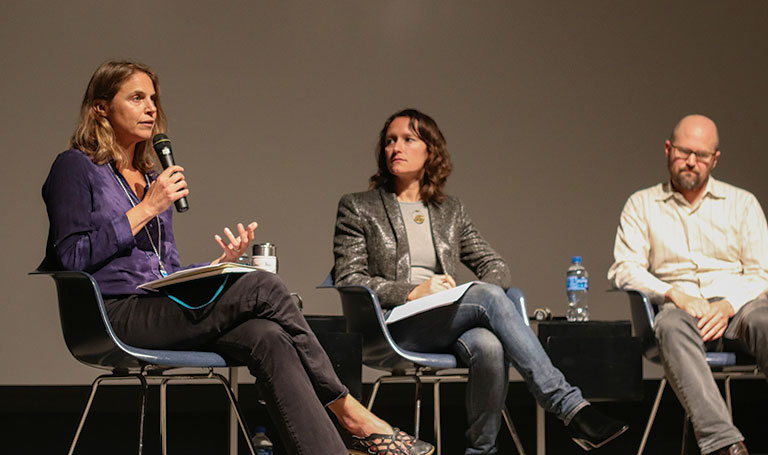 A panel of three speakers engages in discussion at an event, with one woman speaking into a microphone and the others listening attentively.