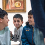 A group of smiling children sit together in an art gallery, sharing laughter and joy, with paintings visible in the background.