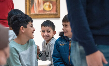 A group of smiling children sit together in an art gallery, sharing laughter and joy, with paintings visible in the background.