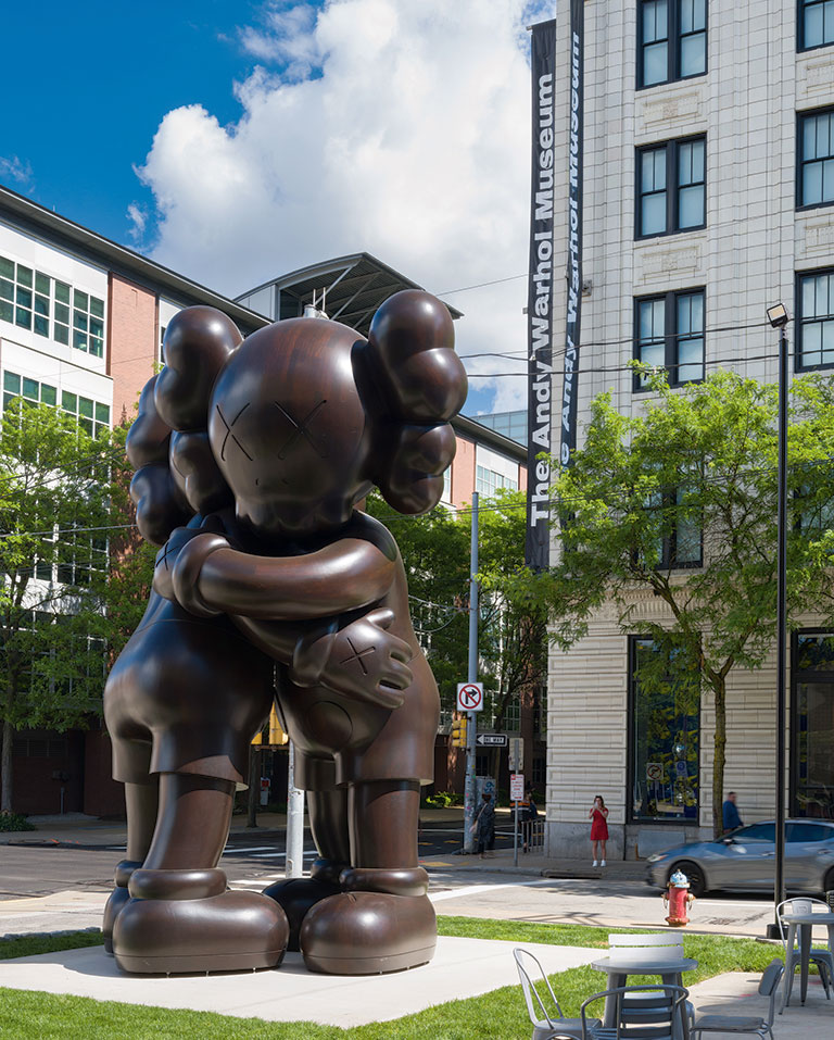 A large bronze sculpture of two cartoon-like figures hugging, located outside the Andy Warhol Museum amidst trees and buildings.