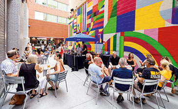 A vibrant outdoor gathering with people seated at tables, colorful mural in the background, and a small serving area.