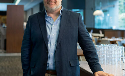 A man in a blazer stands confidently by a bar counter in a modern restaurant, showcasing a relaxed yet professional demeanor.
