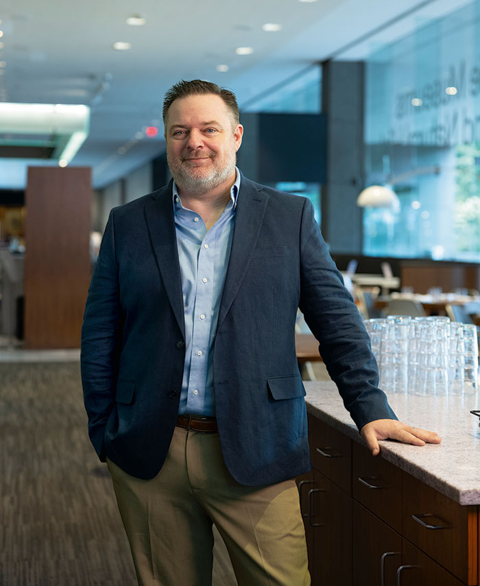 A man in a blazer stands confidently by a bar counter in a modern restaurant, showcasing a relaxed yet professional demeanor.