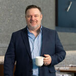 A smiling man in a dark blazer holds a coffee cup, standing in a modern café with a textured wall in the background.
