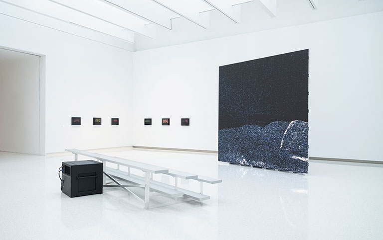 A modern art gallery featuring a large dark wall piece, small framed artworks, and a minimalist bench with a speaker.