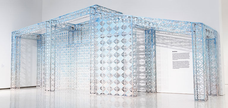 An art installation resembling a translucent structure made of blue and clear panels, with an informational plaque nearby.