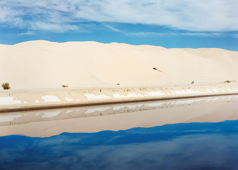 A tranquil desert scene with towering sand dunes under a blue sky, reflecting in a calm water canal below.