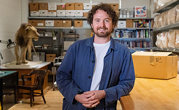A person with curly hair stands in a room filled with shelves of boxes and books, resting hands on a wooden table.