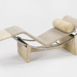 Curved, modern chaise lounge with a chrome frame, beige woven fabric, and a cylindrical pillow. The design is minimalist and elegant.