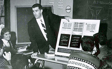 A teacher in a suit passionately gestures at a display board in a classroom. Attentive students sit around, creating an engaged and focused atmosphere.