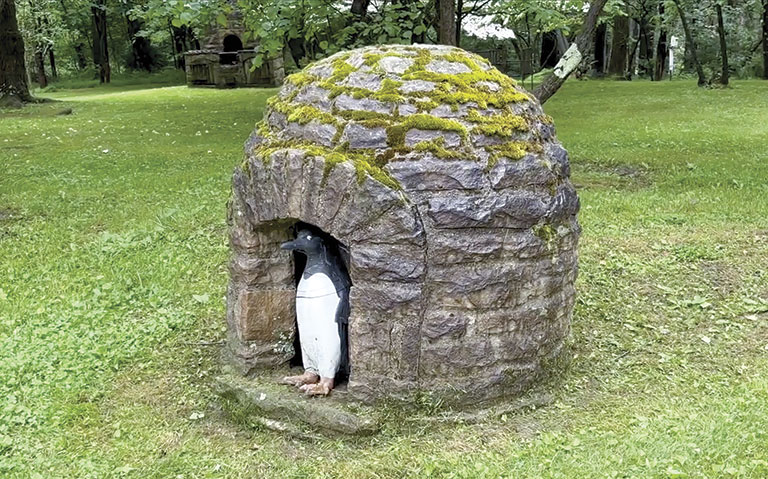 A sculpture of a penguin statue stands inside a moss-covered stone igloo in a lush, green park. The peaceful setting adds a whimsical tone to the scene.