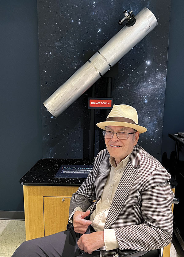 A smiling elderly man in a hat and checkered jacket sits in front of a large telescope with a "Do Not Touch" sign, against a starry backdrop.
