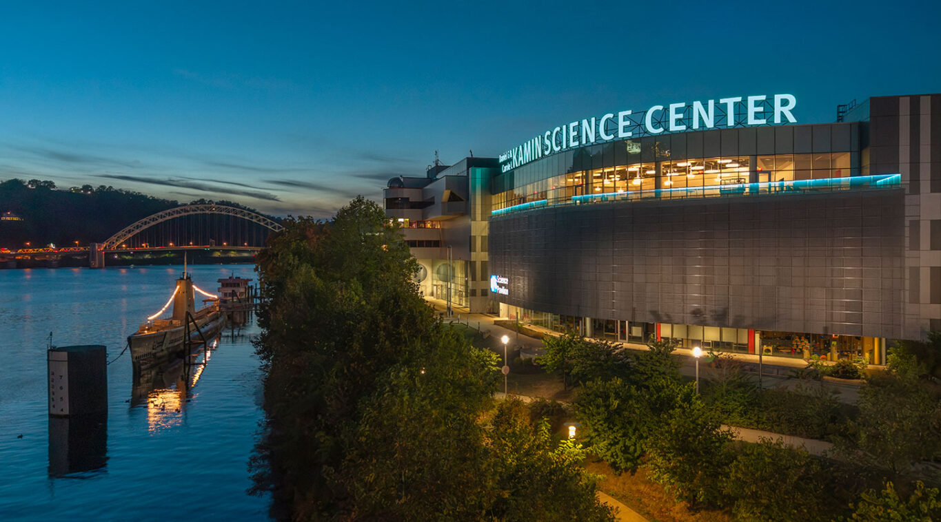 Evening view of a modern science center by a river, with a lit ship docked nearby. A bridge and hills are visible under a twilight sky.
