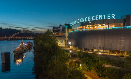 Evening view of a modern science center by a river, with a lit ship docked nearby. A bridge and hills are visible under a twilight sky.