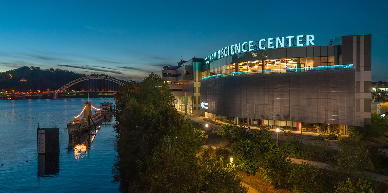Evening view of a modern science center by a river, with a lit ship docked nearby. A bridge and hills are visible under a twilight sky.