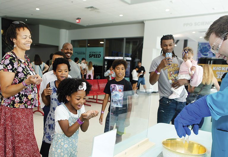 A diverse group of smiling children and adults watch a science experiment. A scientist in safety goggles demonstrates in front of them, creating excitement.