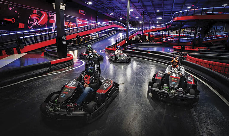 Five people in go-karts race on an indoor track with vibrant lighting and sharp turns. The scene is dynamic and filled with energy and excitement.
