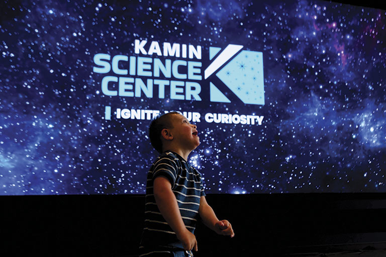 Boy in striped shirt smiles joyfully against a starry backdrop. "Kamin Science Center: Ignite Your Curiosity" is displayed in glowing text above.