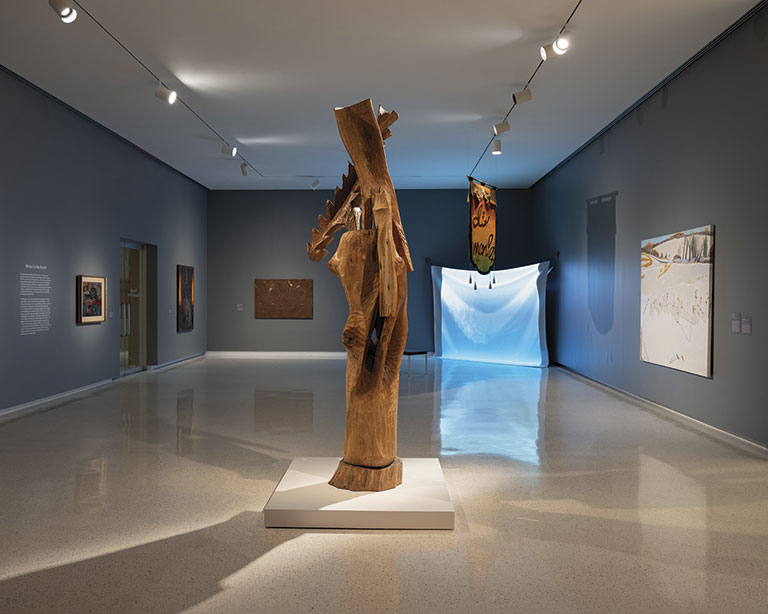 A minimalist art gallery with a tall wooden sculpture as the focal point. The room has smooth gray walls and various abstract paintings, creating a calm ambiance.