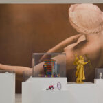 Gallery with large artwork of a reclining figure on the wall. Foreground features modern sculptures on white pedestals, including a golden figure. Traditional paintings frame the scene.