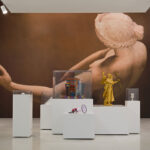 Gallery with large artwork of a reclining figure on the wall. Foreground features modern sculptures on white pedestals, including a golden figure. Traditional paintings frame the scene.