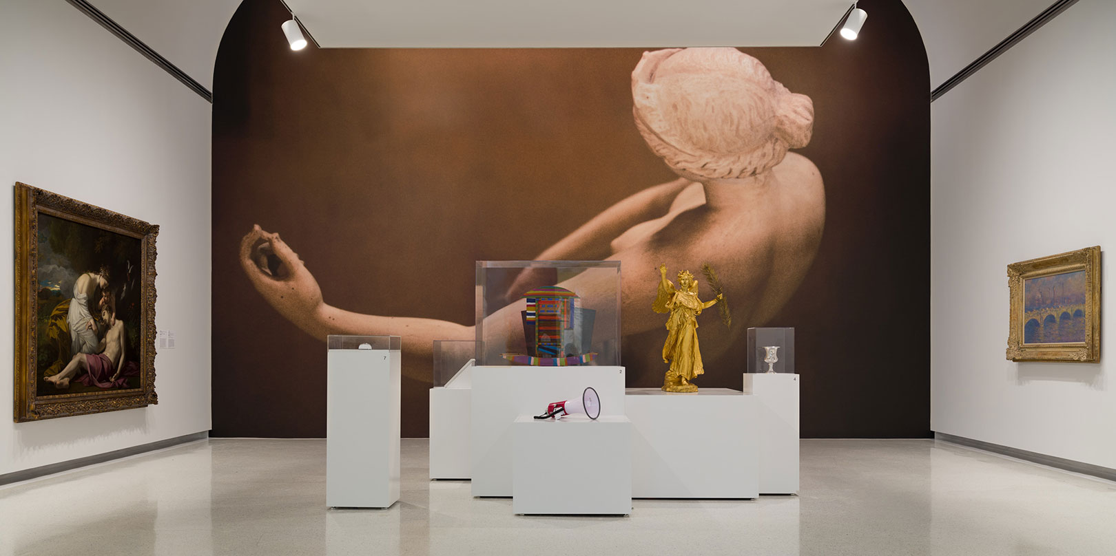 Gallery with large artwork of a reclining figure on the wall. Foreground features modern sculptures on white pedestals, including a golden figure. Traditional paintings frame the scene.