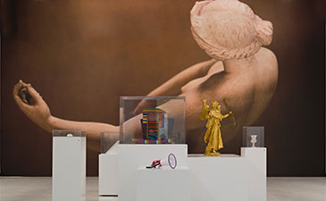 Gallery with large artwork of a reclining figure on the wall. Foreground features modern sculptures on white pedestals, including a golden figure. Traditional paintings frame the scene.