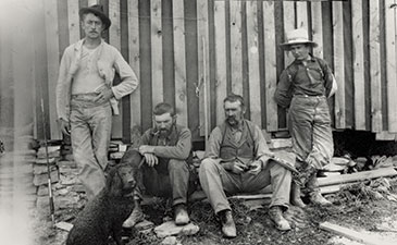 Four men in 19th-century attire and a dog pose by a wooden building. Three are seated on the ground, one stands with arms crossed, creating a relaxed atmosphere.