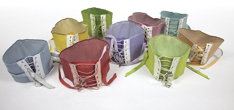 Colorful leather corsets in various hues—red, green, yellow, blue—arranged together. Each features intricate lacing, conveying a sense of craft.